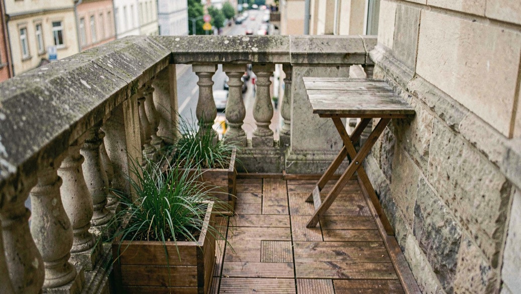 Een smal stadsbalkon met een houten vlonder, twee lage plantenbakken met siergras en een opklapbaar tafeltje tegen een gemetselde balustrade.
